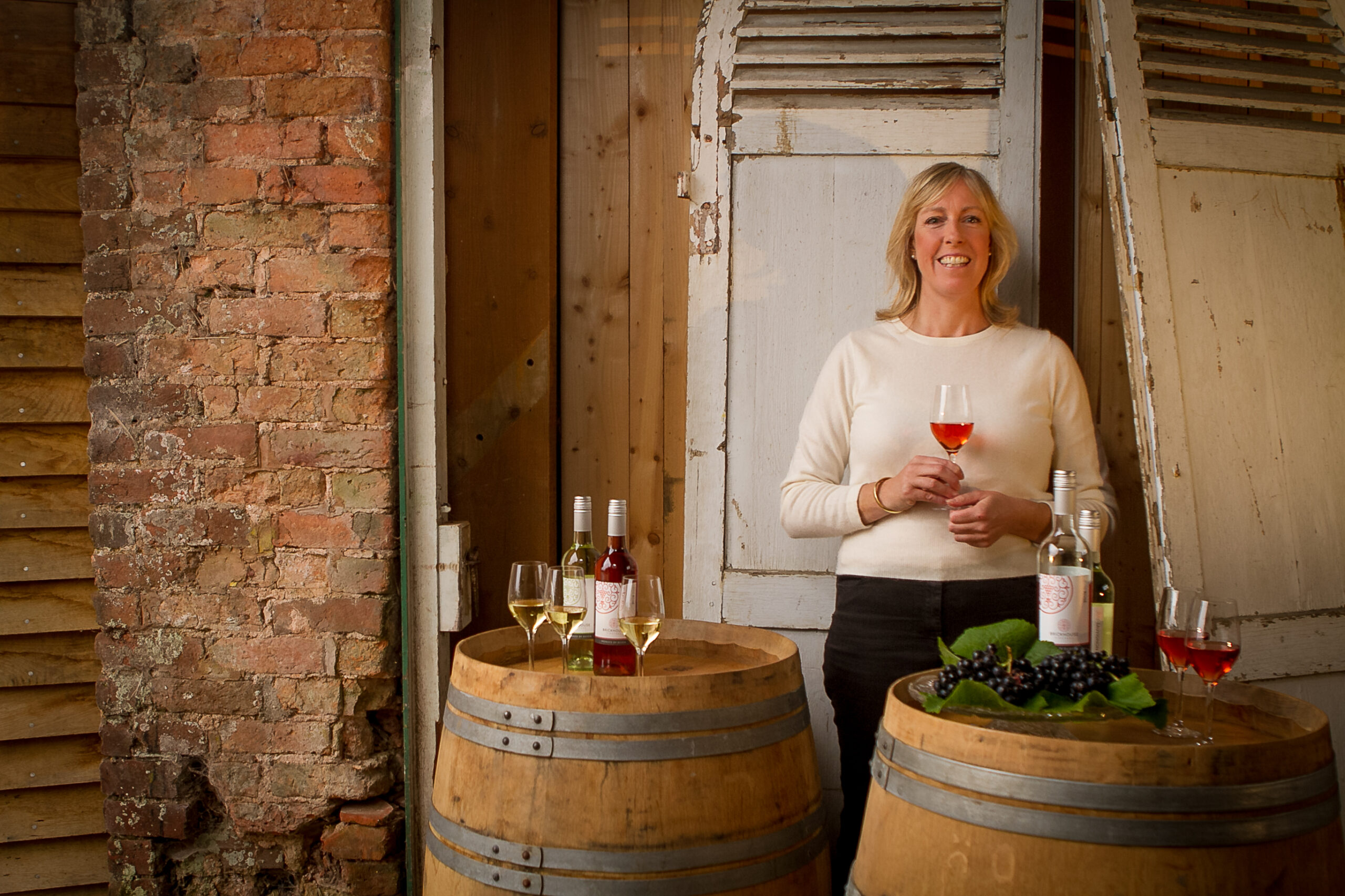 Rebecca Mitchell - Tasting Host is stood behind 2 wine barrels with glasses and bottles of wine on top. Against a rustic brick background.