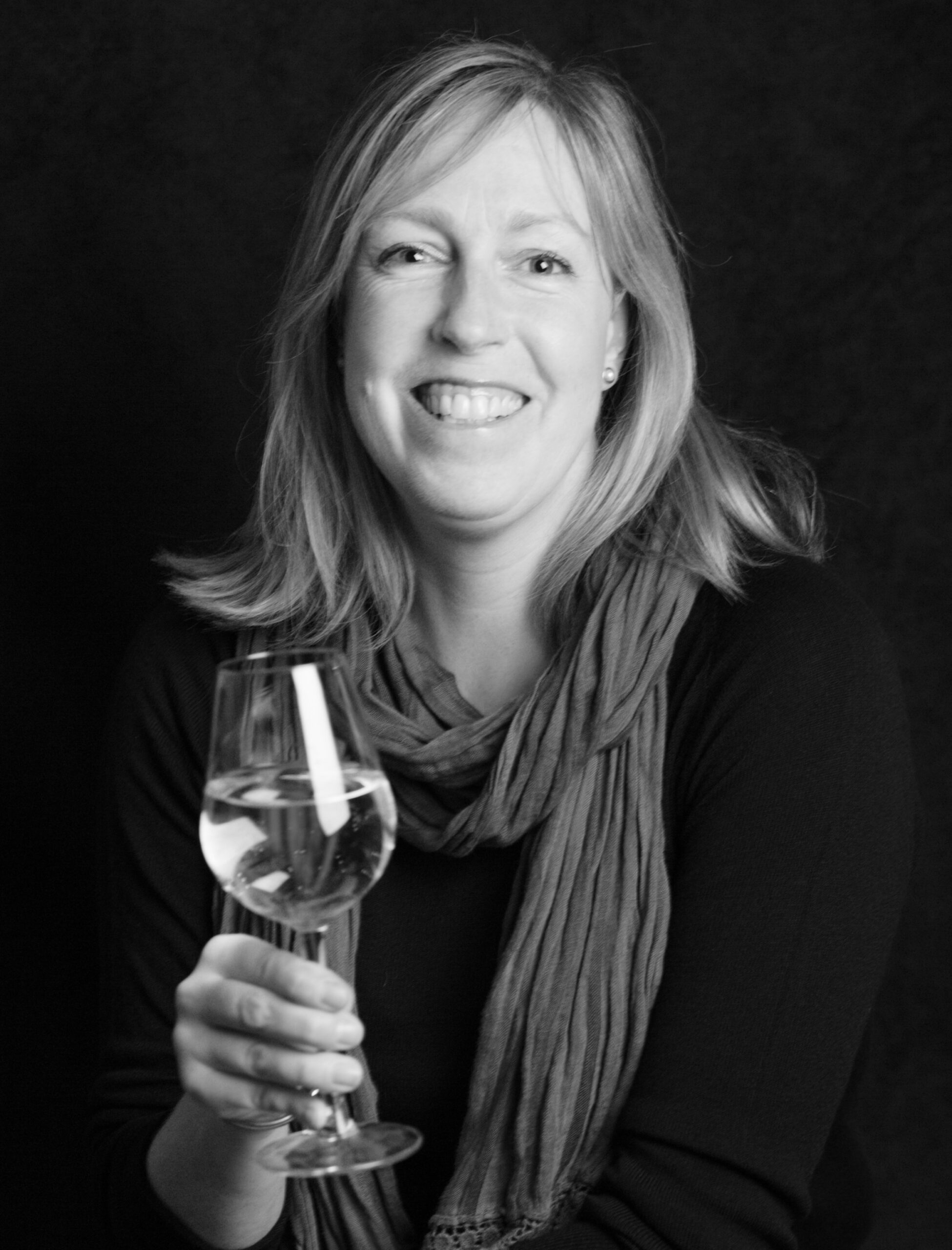 black and white image of Rebecca Mitchell with long hair, wearing a scarf and dark top, holding a glass of wine.